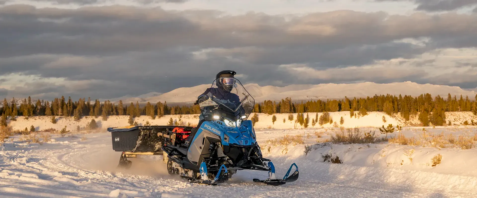 Rider on a blue Polaris RMK Factory Edition towing a cargo sled through snow at sunset