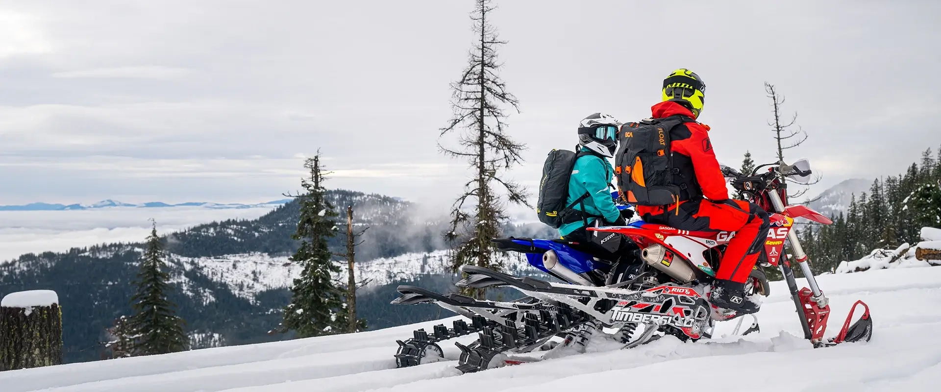 Riders on red and blue snow bikes with Timbersled kits on a snowy mountain ridge overlooking a cloud-filled valley