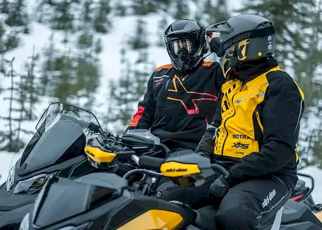 Two riders on snowmobiles in a snowy forest, one wearing a black and red jacket and the other in yellow and black