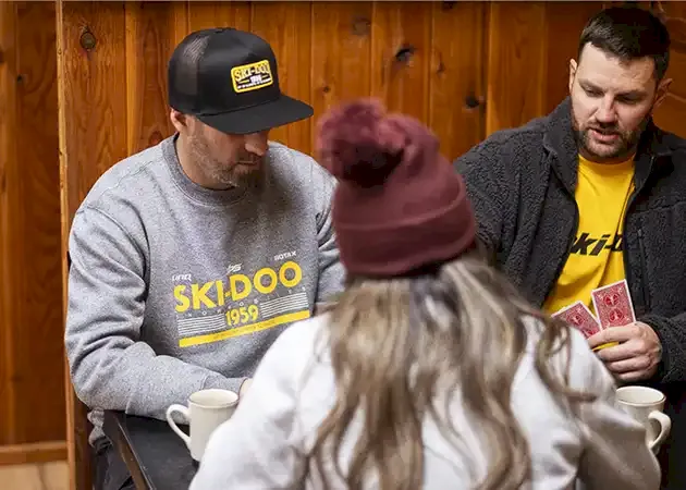 Two men and a woman sitting around a table playing cards inside a wood-paneled cabin