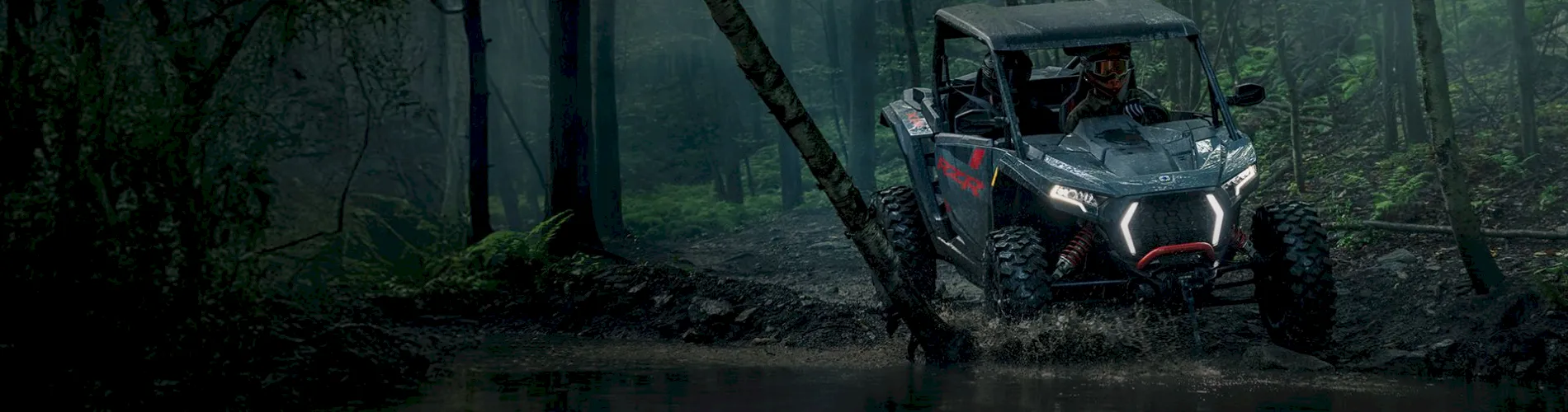 Polaris RZR UTV splashing through a muddy stream in a dense misty forest