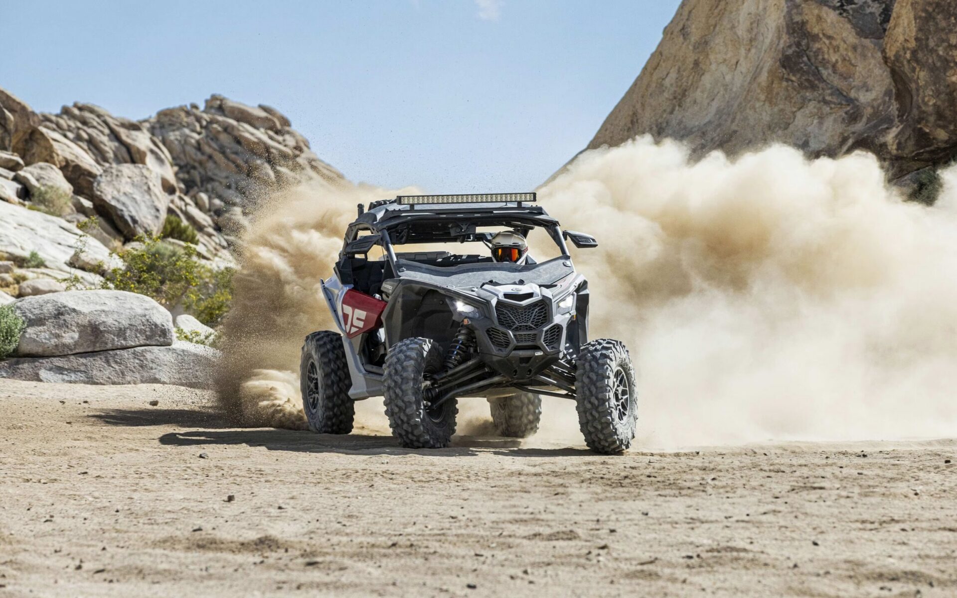 Rider on a dark olive ATV navigating a steep, rocky mountain ridge under a clear sky with other riders following behind